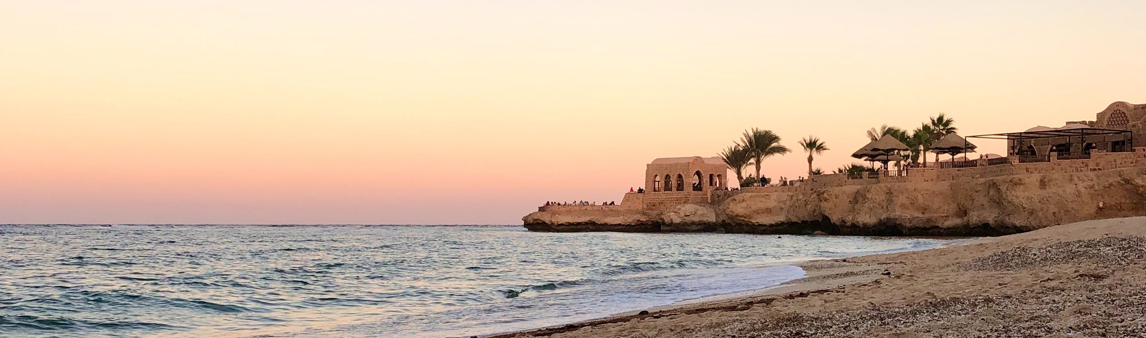 Sunset view of Movenpick Resort El Quseir at Sirena Beach on Egypt’s Red Sea coast, featuring calm waves, a sandy shore, and traditional stone architecture with palm trees on a rocky cliff.