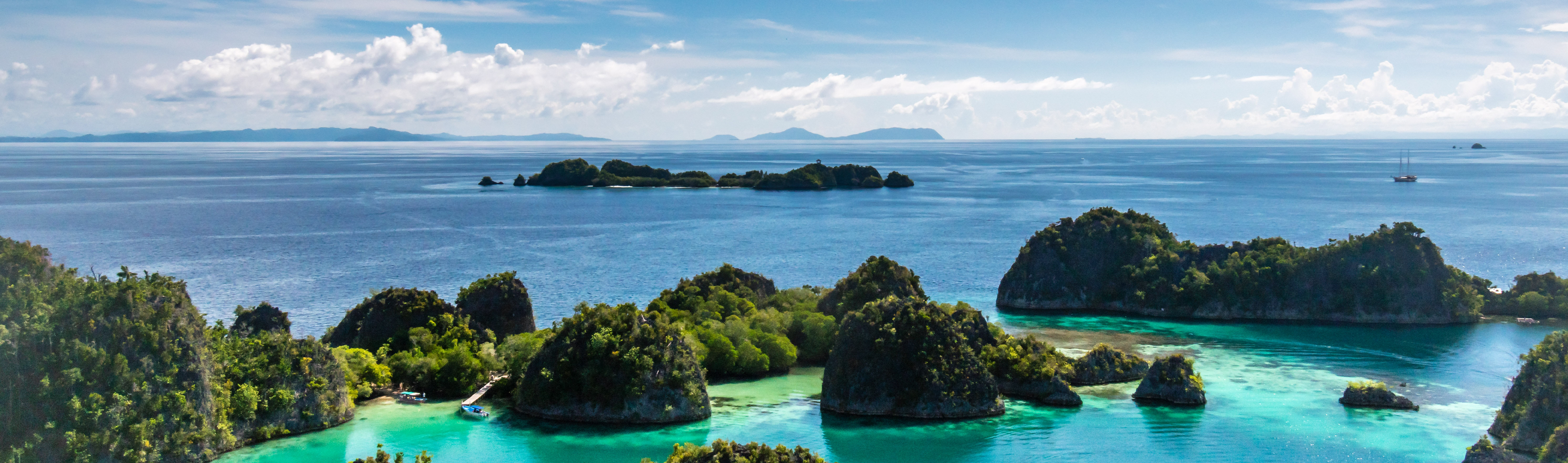 Aerial view of the lush green islets and clear turquoise waters of Raja Ampat, Indonesia, with distant islands and a sailing yacht on the calm ocean horizon under a partly cloudy sky.