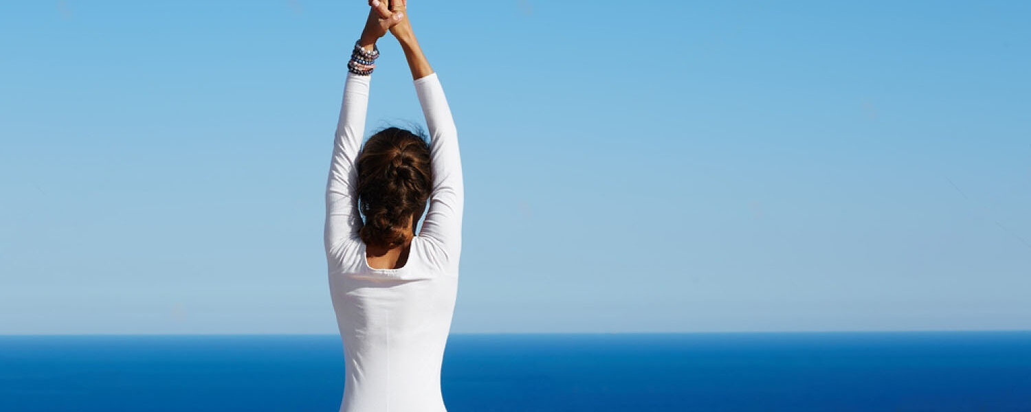 A woman is doing a yoga pose outdoors with the sea and a blue sky in the background